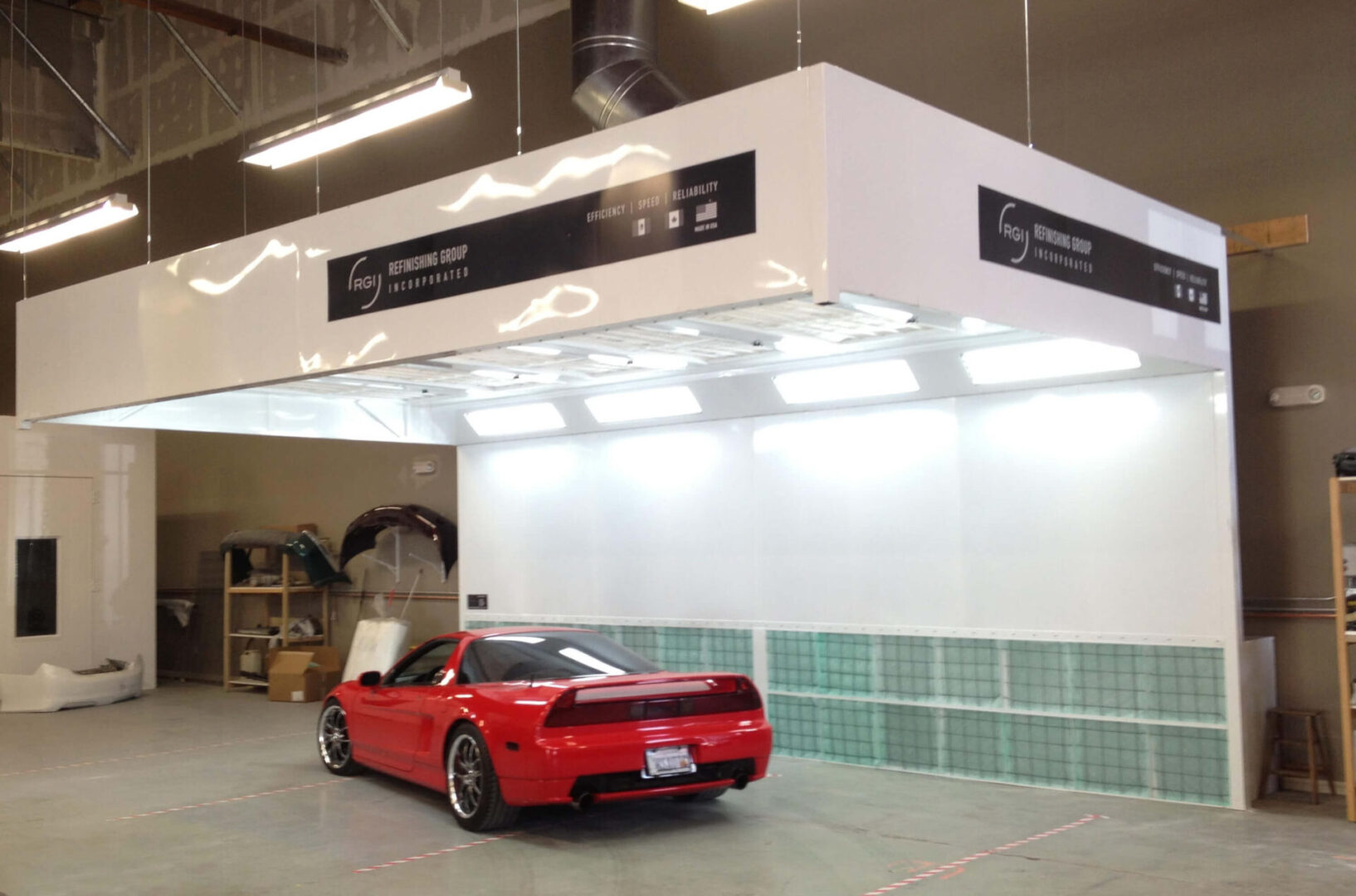 A red sports car parked under a large white industrial booth with illuminated panels and signage displaying "fog retention system" in a warehouse setting.