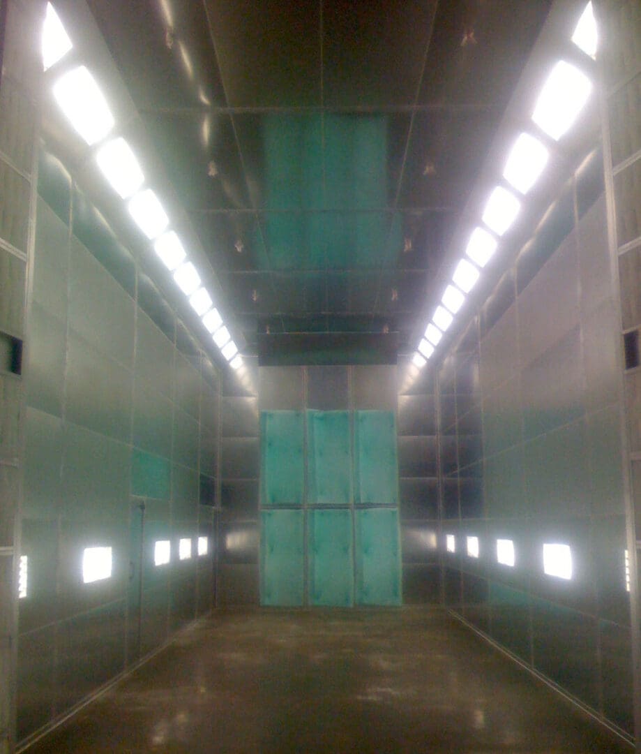 Interior of an industrial empty room with shiny metal walls and ceiling, lit by multiple lights, featuring a large teal panel at the far end.