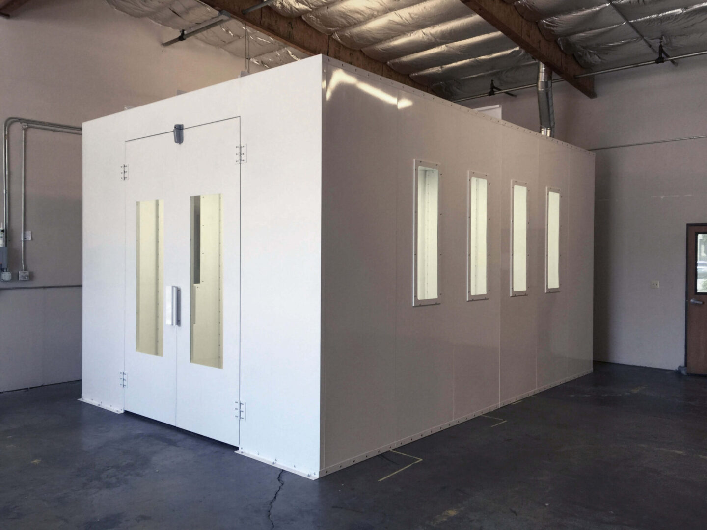 A large, white modular structure with a door and multiple small windows, situated in an empty room with exposed beams and concrete flooring.