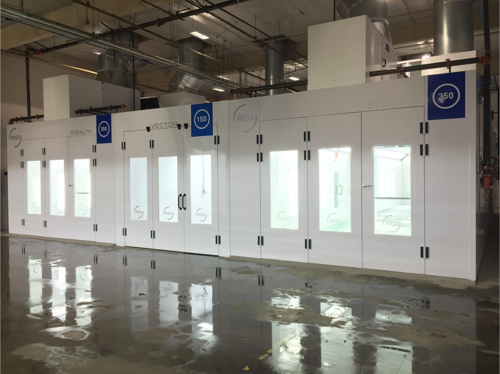 Industrial spray booths in a large workshop, featuring white exteriors with glass doors and company logos, and a reflective wet floor.