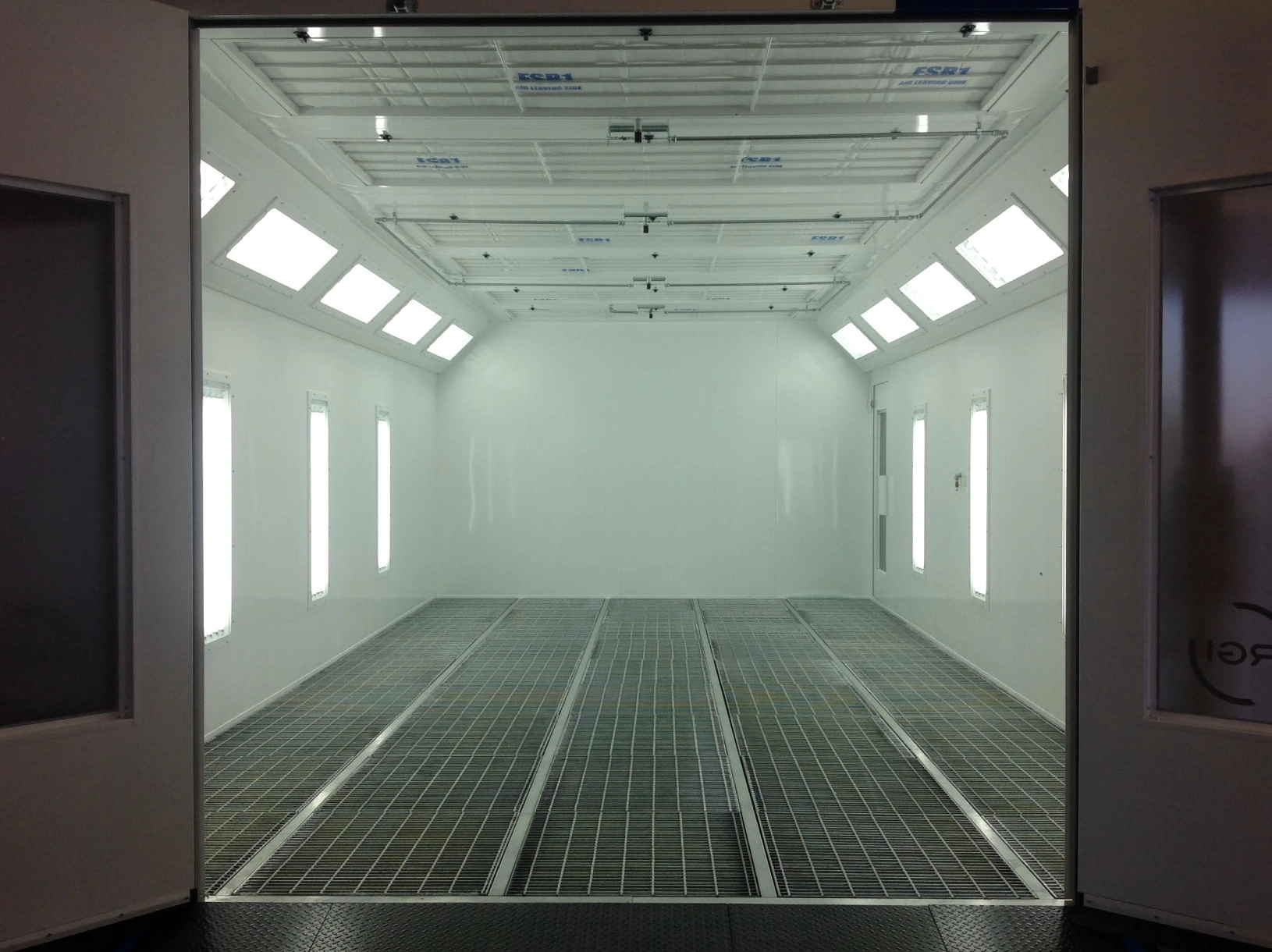 Empty industrial room with a grid floor, overhead lights, and large glass doors.